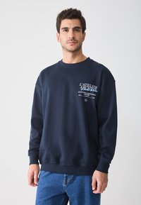Marineblaue Sweatshirt mit Rundhalsausschnitt, gerippten Bündchen und Saum. Auf der Vorderseite mit weißem Drucktext versehen. Kombiniert mit blauen Jeans.