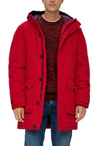 Parka rossa imbottita con cappuccio, dotata di bottoni automatici, due tasche frontali e una foderatura interna texturizzata. Indossata sopra un maglione a motivo rosso.