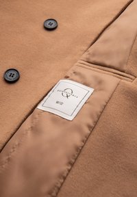 Close-up af beige uldfrakke med to mørke knapper og en indvendig satinforing med en hvid etiket med teksten "Quintessence M 48."
