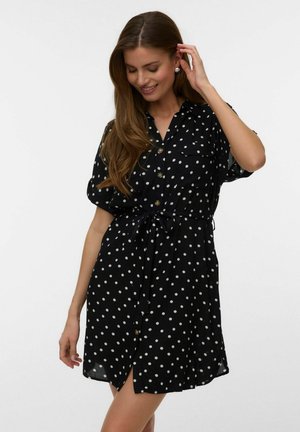 Vrouw glimlachend, draagt een zwarte korte mouw blousejurk met witte polkadots en een ceintuur in de taille, terwijl ze haar haar aanraakt.