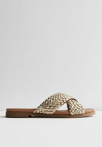 Kudotun kultaisenvärinen sandaali, jossa on ristitäännykset, tasainen pohja ja sileä ruskea pohjallinen. Yksinkertainen muotoilu ilman lisäkoristuksia.