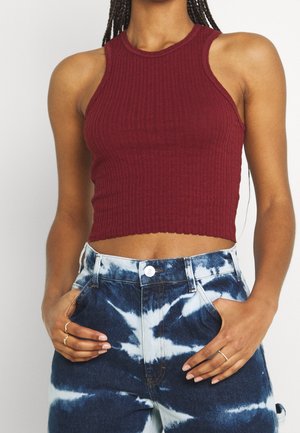 Femme portant un crop top bordeaux sans manches, côtelé, et un jean taille haute bleu et blanc tie and dye, les mains dans les poches.