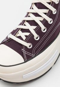 Fioletowy trampki na płóciennym materiale z białymi sznurówkami, gumowym czubkiem i teksturowaną, czarną podeszwą. Posiada srebrne oczka i kontrastujące przeszycia.