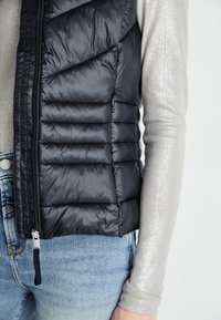 Gilet noir rembourré avec un design matelassé, fermeture éclair et poches latérales. Porté par-dessus un haut argenté à manches longues et un jean bleu.