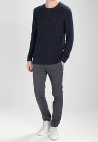 Pull en maille torsadée de couleur marine avec encolure ras du cou, associé à un pantalon gris slim et des baskets blanches. Le pull arbore un logo de marque discret.