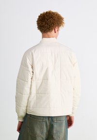 Lys beige quiltet jakke med en kasseformet silhuet, ribbede manchetter og en kort opretstående krave, parret med falmede denimbukser.