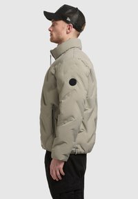 Uomo che indossa un piumino beige con colletto alto e tasche con zip nere, cappellino da baseball in rete nera, pantaloni cargo neri, di profilo rivolto verso sinistra.