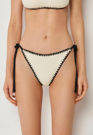 Kvinde iført hvidt tekstureret bikinitrusser med sort zigzagkant og sidelåse, stående mod en neutral baggrund.