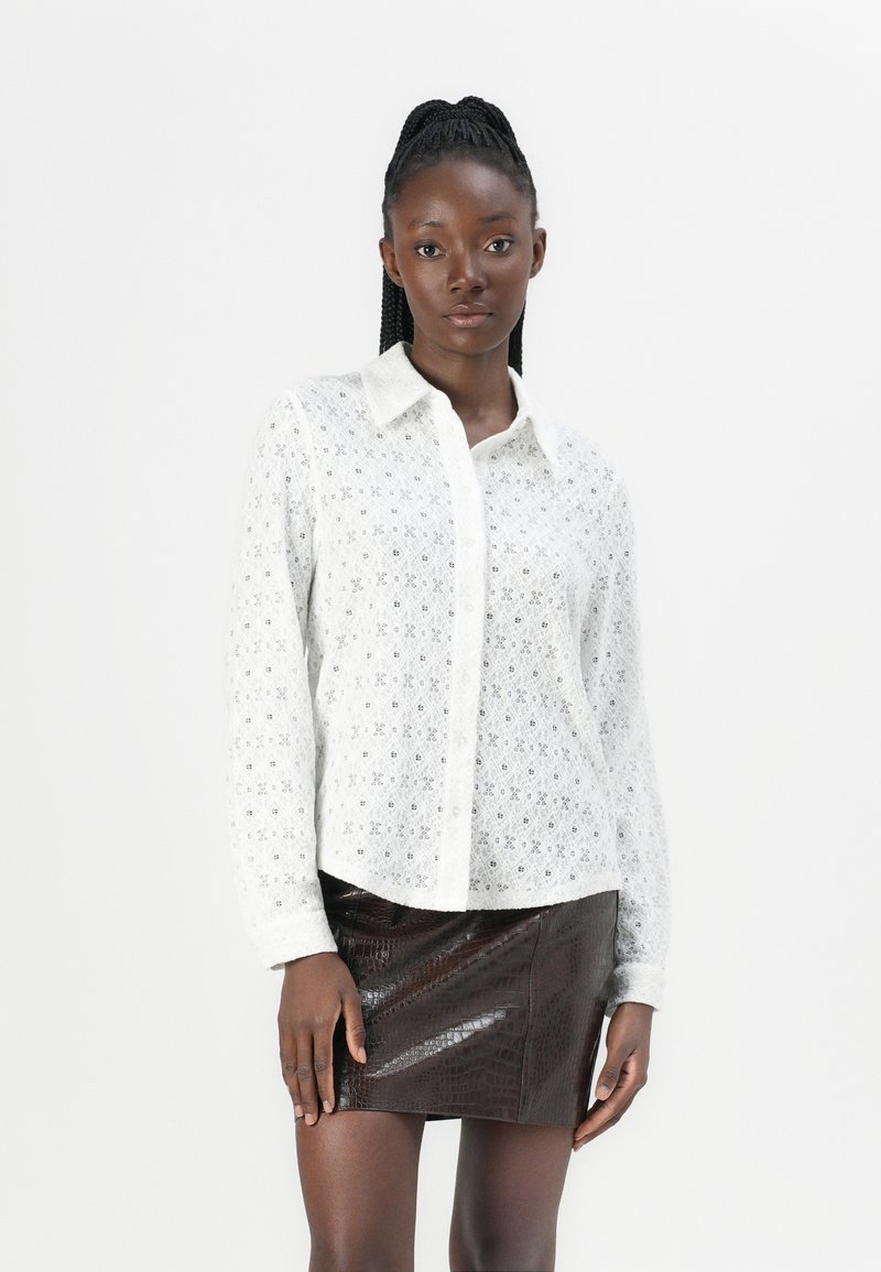 Blusa bianca in pizzo con colletto, chiusura a bottoni e maniche lunghe. Indossata con una mini gonna in leather marrone con texture. Tessuto liscio e leggero.