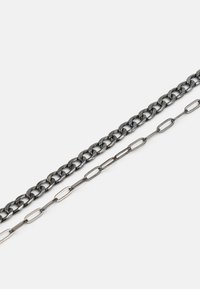 Duas correntes: uma grossa, entrelaçada, de metal escuro com um acabamento brilhante; a outra uma corrente mais fina e alongada com um aspeto mate.