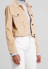 Veste cropped en velours côtelé beige avec deux poches poitrine, fermeture à bouton et texture côtelée. Associée à un col roulé gris et un jean blanc.