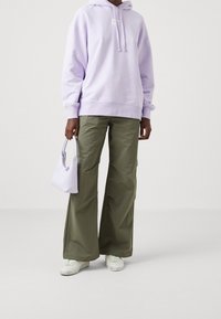 Felpa oversized viola, pantaloni a gamba larga verde oliva, sneakers bianchi e una piccola borsa lavanda. Texture morbide e design informale.