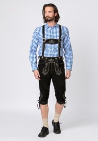 Black suede lederhosen with silver embroidery, paired with a blue checkered shirt. Features shoulder straps and knee-length design. Black shoes and light socks.