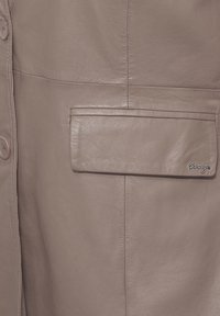 Leren jack in zacht taupe, met knoopsluitingen, een platte zak met een afgeronde rand en een subtiel merkembleem. Gladde textuur.