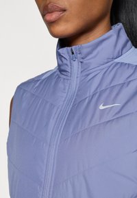 Vest mov cu mânecă scurtă, cu guler înalt, design matlasat și închidere cu fermoar. Prezintă un mic logo alb Nike pe piept. Textură netedă.