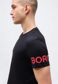 Άνδρας με μαύρο πουκάμισο κοντομάνικο με κόκκινα διάτρητα γράμματα "BORG" στο αριστερό μανίκι, κοιτάζει προς τα αριστερά μπροστά σε απλό φωτεινό φόντο.