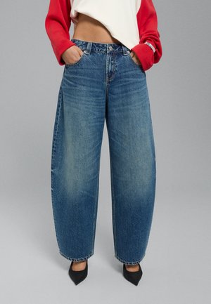 Personne portant un jean en denim bleu ample, des chaussures noires pointues, un top blanc court et un vêtement rouge à manches longues avec les mains dans les poches.