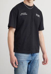 Camiseta de algodón negra con mangas cortas, cuello redondo y estampado en blanco que dice "LOS FUMADORES MUEREN JÓVENES" en el frente, con el logo de "PEIDUS".