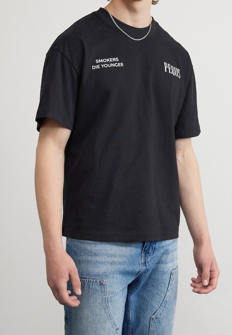 Camiseta de algodón negra con mangas cortas, cuello redondo y estampado en blanco que dice "LOS FUMADORES MUEREN JÓVENES" en el frente, con el logo de "PEIDUS".