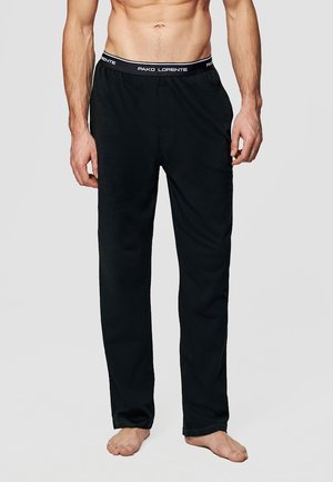 Mężczyzna w czarnych spodniach o luźnym kroju z elastycznym pasem i białym logo "PAKO LORENTE", stojący boso na jednolitym tle.