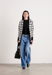 Cardigan com padrão preto e branco, blusa preta, jeans azuis largos e sapatos pretos de bico apontado; segurando uma bolsa preta; texturas suaves.