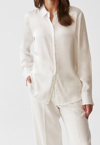 Vit satin långärmad skjorta med knappstängning fram, klassisk krage och avslappnad passform. Tyget har en slät, glansig yta.