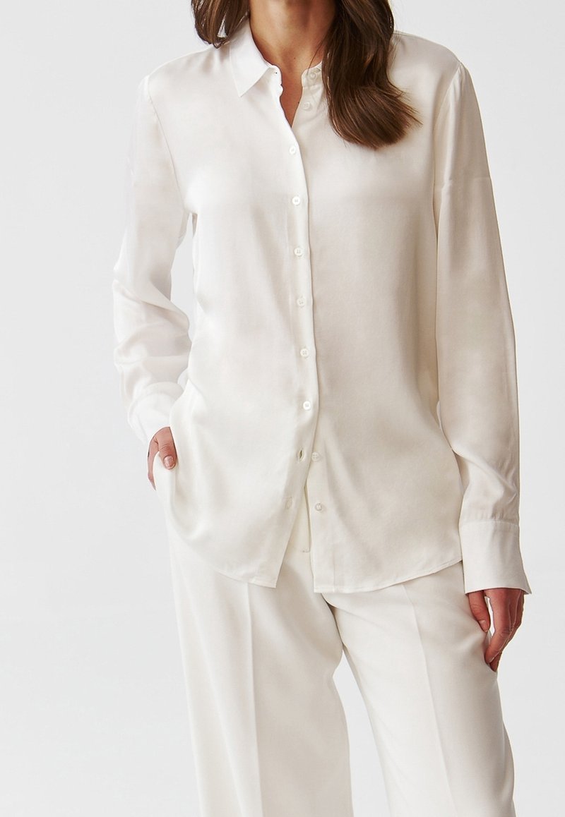 Vit satin långärmad skjorta med knappstängning fram, klassisk krage och avslappnad passform. Tyget har en slät, glansig yta.