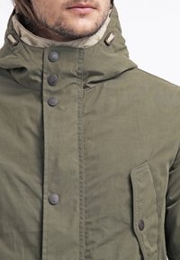 Olive green jacket with a hood, featuring a beige lining, multiple front buttons, textured fabric, and a side pocket.