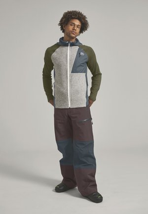 Mann mit lockigem Haar, der einen grünen und grauen Kapuzenpullover mit Reißverschluss, lockere braune Hosen mit blauen Flicken und schwarze Schuhe trägt, steht vor einem einfarbigen Hintergrund.