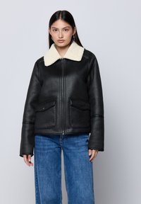 Giacca in ecopelle nera con colletto in shearling crema, chiusura con zip e due tasche frontali. Presenta una vestibilità strutturata e maniche ampie.