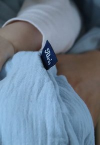 Helles blaues Gewebe wird gehalten und zeigt eine strukturierte Oberfläche. Ein dunkelblaues Etikett ist angebracht, auf dem "Alvi" in weißer Schrift zu sehen ist.