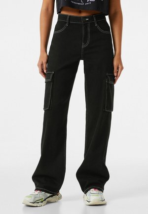 Persona que lleva pantalones cargo de pierna ancha negros con grandes bolsillos laterales y costuras blancas, combinados con zapatillas multicolores.