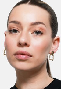 Goldfarbene Ohrringe mit texturiertem Design, die eine unregelmäßige rechteckige Form und eine gebogene Akzentuierung an der Oberseite aufweisen, getragen von einem Model.