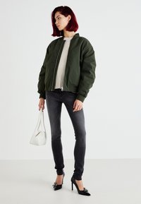Chaqueta bomber verde, suéter beige claro, jeans ajustados grises, tacones altos negros y un pequeño bolso blanco. Corte relajado con cuello estilo stand.