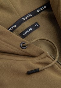 Braune Kapuzenjacke mit schwarzer Kordel und Reißverschluss, mit den Markenlabels „ombre“ und „basic“ im Krageninnenbereich.