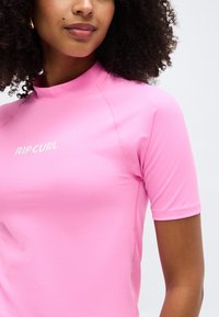 Γυναίκα με σγουρά μαλλιά που φοράει φωτεινό ροζ κοντομάνικο μπλουζάκι προστασίας από τον ήλιο με το λογότυπο «RIP CURL» στο στήθος.