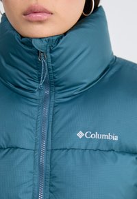 Tyrkisk nedjakke med høy krage, full glidelås og Columbia-logo. Har quiltet design og jevn tekstur.