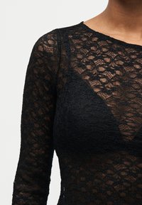 Γυναίκα που φοράει μακρυμάνικη μαύρη δαντελένια μπλούζα με λουλουδάτα μοτίβα και από κάτω ένα μαύρο δαντελένιο bralette που ταιριάζει.