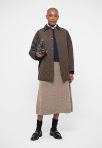 Frau in einer braunen gesteppten Jacke, beigefarbenem gestrickten Midikleid, schwarzen Loafers mit Socken, die eine schwarze Handtasche mit Nieten hält.