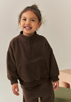 Lille barn smiler, iført en brun fleece-trøje med lynlås og matchende bukser, står indenfor mod en lys baggrund.