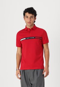 Rød polo shirt med en sort og hvid vandret stribe med "TOMMY HILFIGER" tekst, korte ærmer og en klassisk krave. Grå bukser.