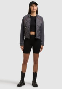 Femme portant une casquette noire, une veste aviateur matelassée grise, un crop top noir, un short de cycliste noir et des bottes noires mi-mollet, debout devant un fond uni.