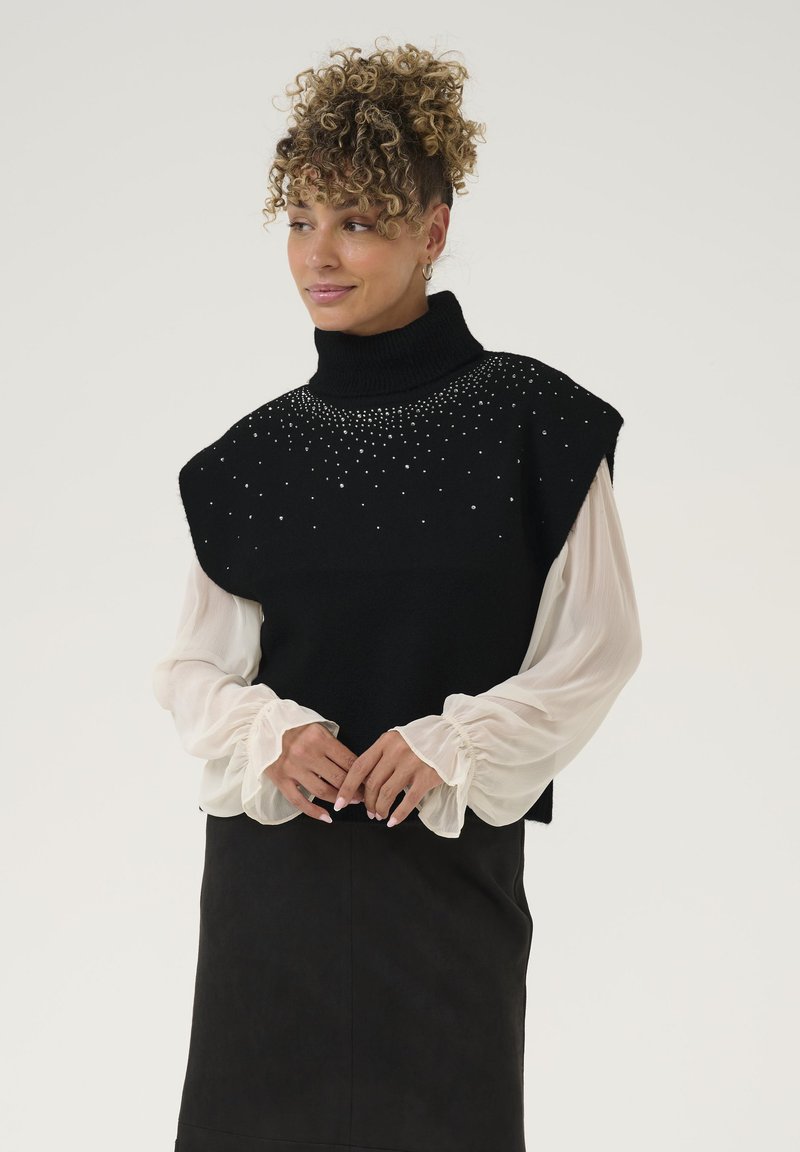 Črn brezrokavnik s turtleneck izrezom, okrašen s perlicami okoli vratu, prekrit z prosojno belo srajco z dolgimi rokavi, v kombinaciji s črno krilom.