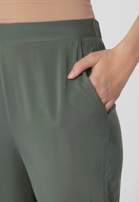Pantaloni leggeri di un verde scuro con una texture liscia, dotati di una tasca laterale e una vita aderente.