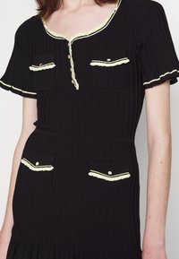 Robe en maille côtelée noire avec manches courtes, quatre poches boutonnées bordées de jaune pâle, et un col et des bords de manches volantés.