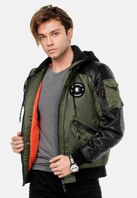 Olivgrüne Bomberjacke mit schwarzen Leder-Schultern, orangefarbenem Innenfutter und mehreren Taschen. Mit Logo-Patch und Reißverschlussakzenten.