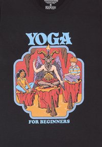Camiseta negra con un gráfico colorido de una figura con cabeza de cabra sentada en una roca, flanqueada por dos niños, con el texto "YOGA PARA PRINCIPIANTES".