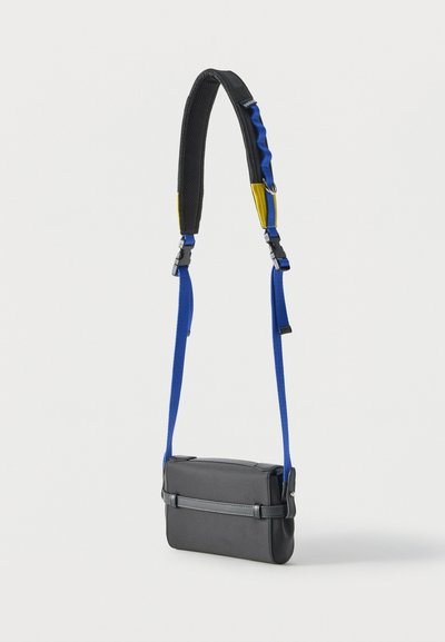 Sac bandoulière rectangulaire noir avec une sangle d'épaule rembourrée ajustable noire, bleue et jaune, suspendue sur un fond blanc.