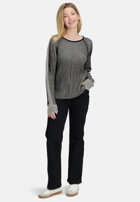 Kobieta uśmiechająca się, w czarno-białym, prążkowanym swetrze z długim rękawem, czarnych spodniach i białych sneakersach, stojąca z jedną ręką w kieszeni.