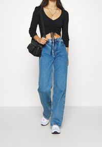 Schwarzes, langärmliges Cropped-Top mit Raffung, blaue weit geschnittene Jeans und weiße Sneaker. Schwarze Umhängetasche mit Klappenverschluss.